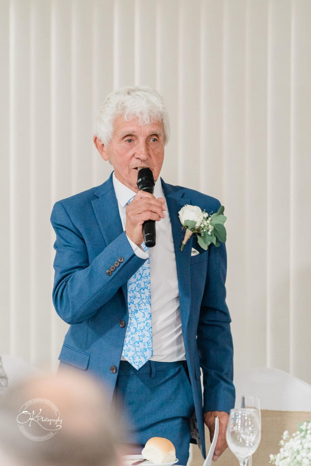 A man in a blue suit with a patterned blue tie is speaking into a microphone at an event.