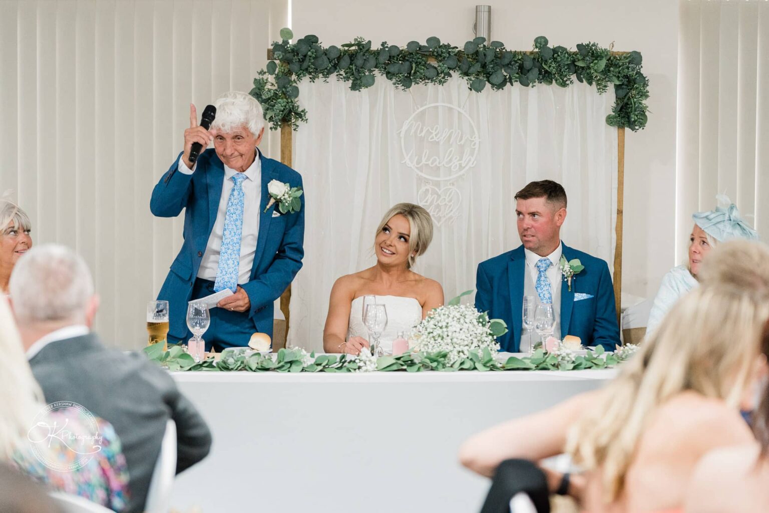 An elderly man in a blue suit gives a speech at a wedding reception, while the bride in a white dress and the groom in a blue suit listen attentively at the head table decorated with greenery and white flowers.