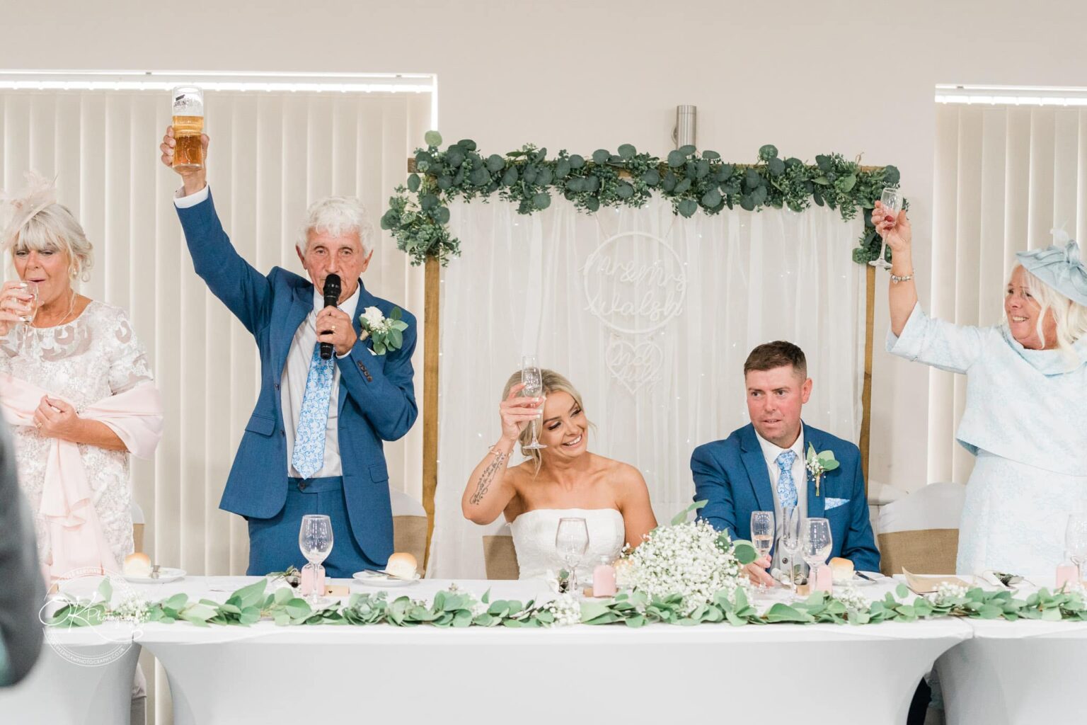 Wedding party raising glasses for a toast, with bride and groom seated at a decorated table.