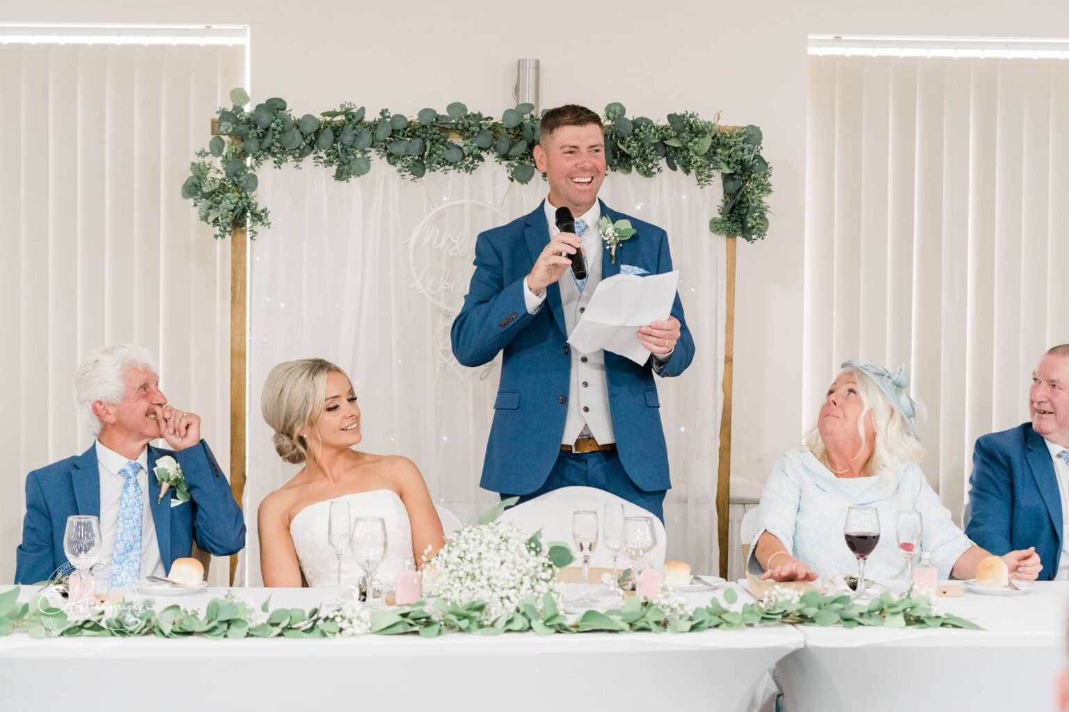 A man in a blue suit gives a speech at a wedding reception while holding a microphone and a piece of paper, with four seated guests, including the bride, listening and smiling.