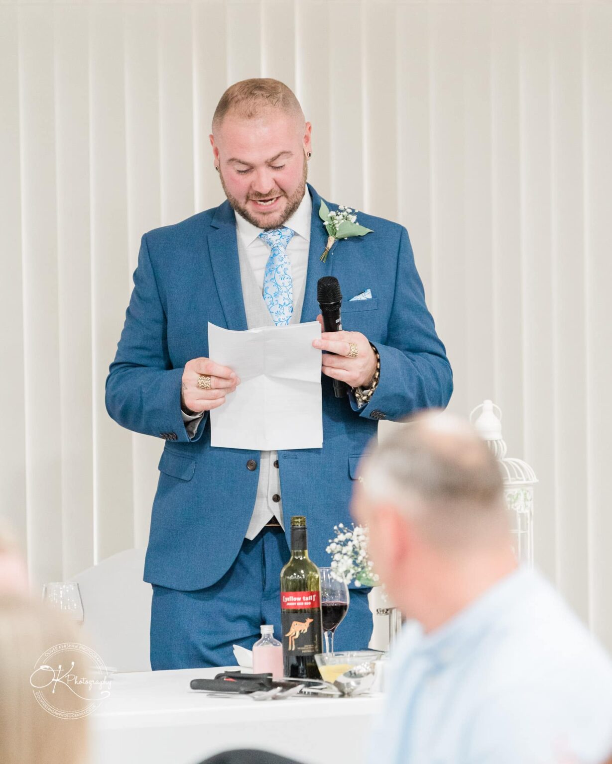 A man in a blue suit gives a speech, holding a microphone and a piece of paper, standing at a table with a wine bottle and a glass.