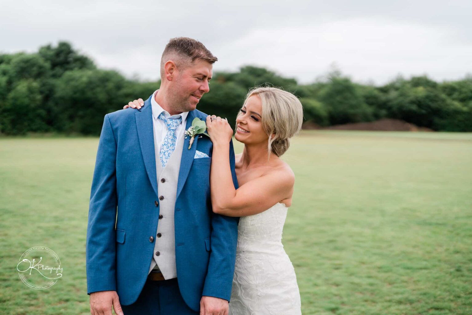 A bride and groom standing on a grassy field, with the bride smiling and leaning on the groom's shoulder.