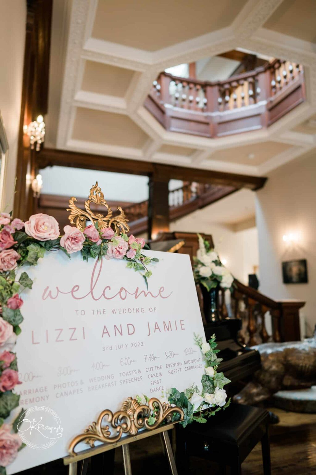 Welcome sign at a wedding with pink flowers, a golden frame, and itinerary details, set in a venue with a wooden staircase and ornate ceiling.