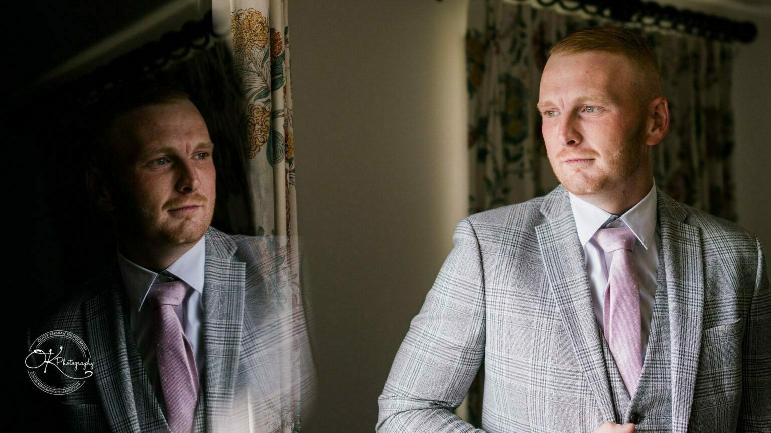 A man in a grey checkered suit with a pink tie looks out a window with floral curtains, his reflection visible on the glass.
