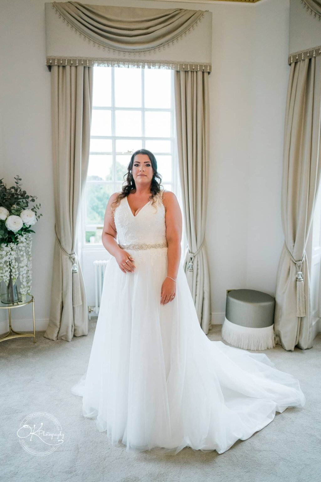 Bride in white wedding dress standing in a room with large window and elegant decor.