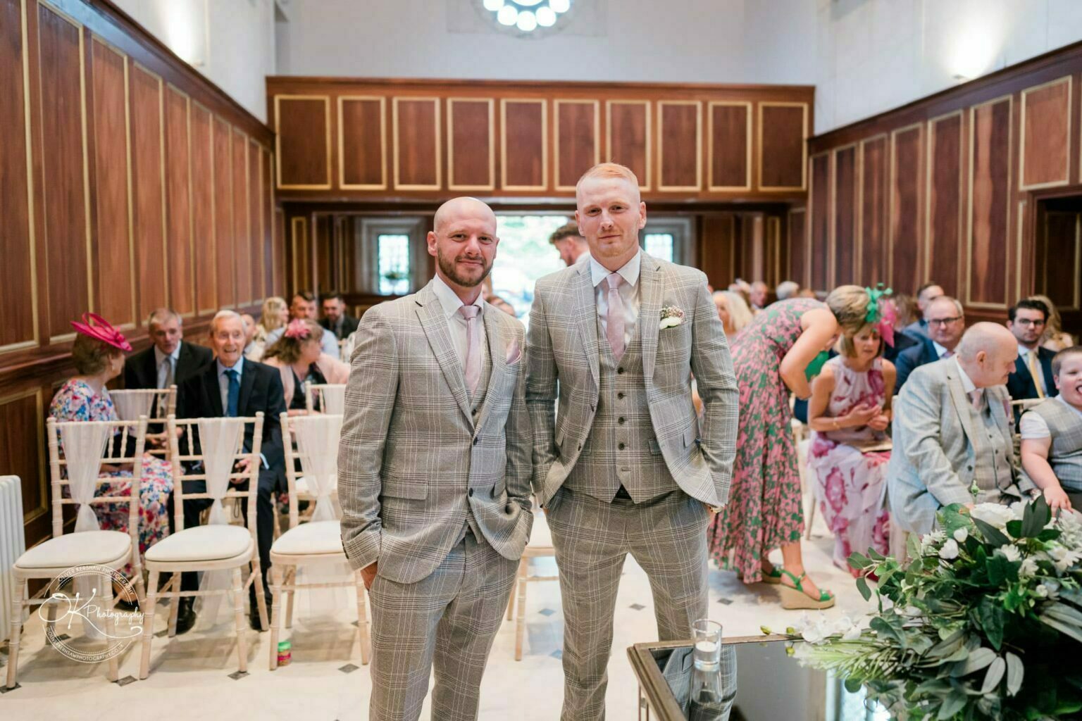 Two men in matching grey plaid suits pose in the foreground of a wedding hall with seated guests at Bourton Hall.