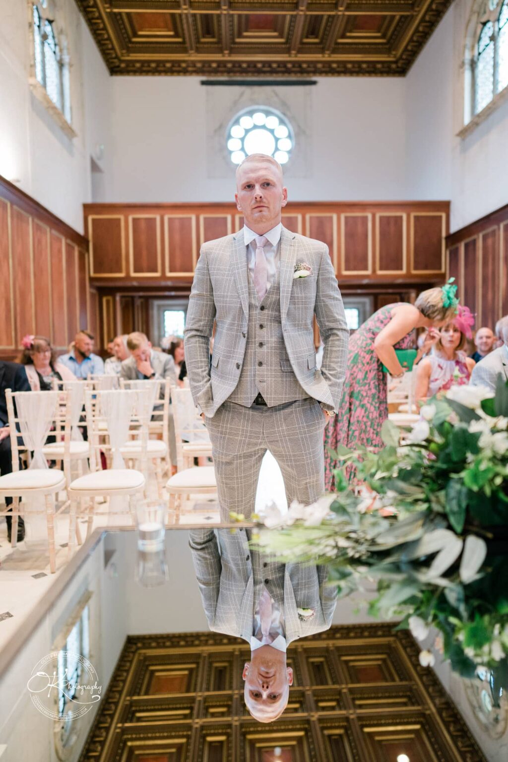 A groom in a checked suit stands confidently in an ornate room with wood paneling and a decorative ceiling, with his reflection visible on a shiny surface in the foreground. Guests are seated in the background.