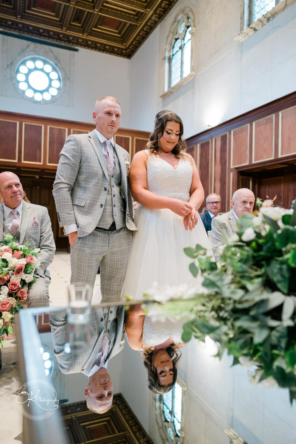 A couple stands together during their wedding ceremony in a grand hall with wooden panelling and arched windows.