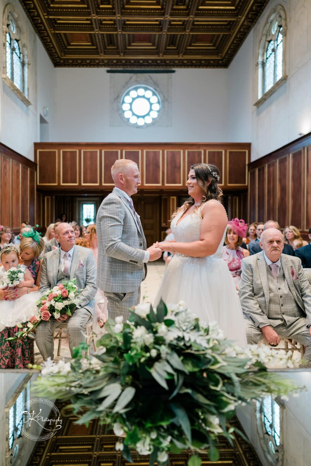A couple exchanging wedding vows at Bourton Hall, surrounded by guests seated in a stylish wooden-panelled interior with elaborate ceiling details.