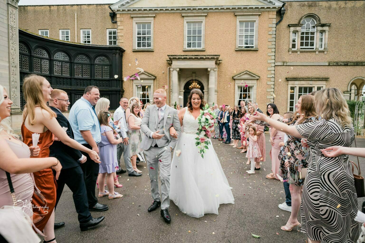 Bride and groom walking down an aisle surrounded by guests throwing confetti outside Bourton Hall.