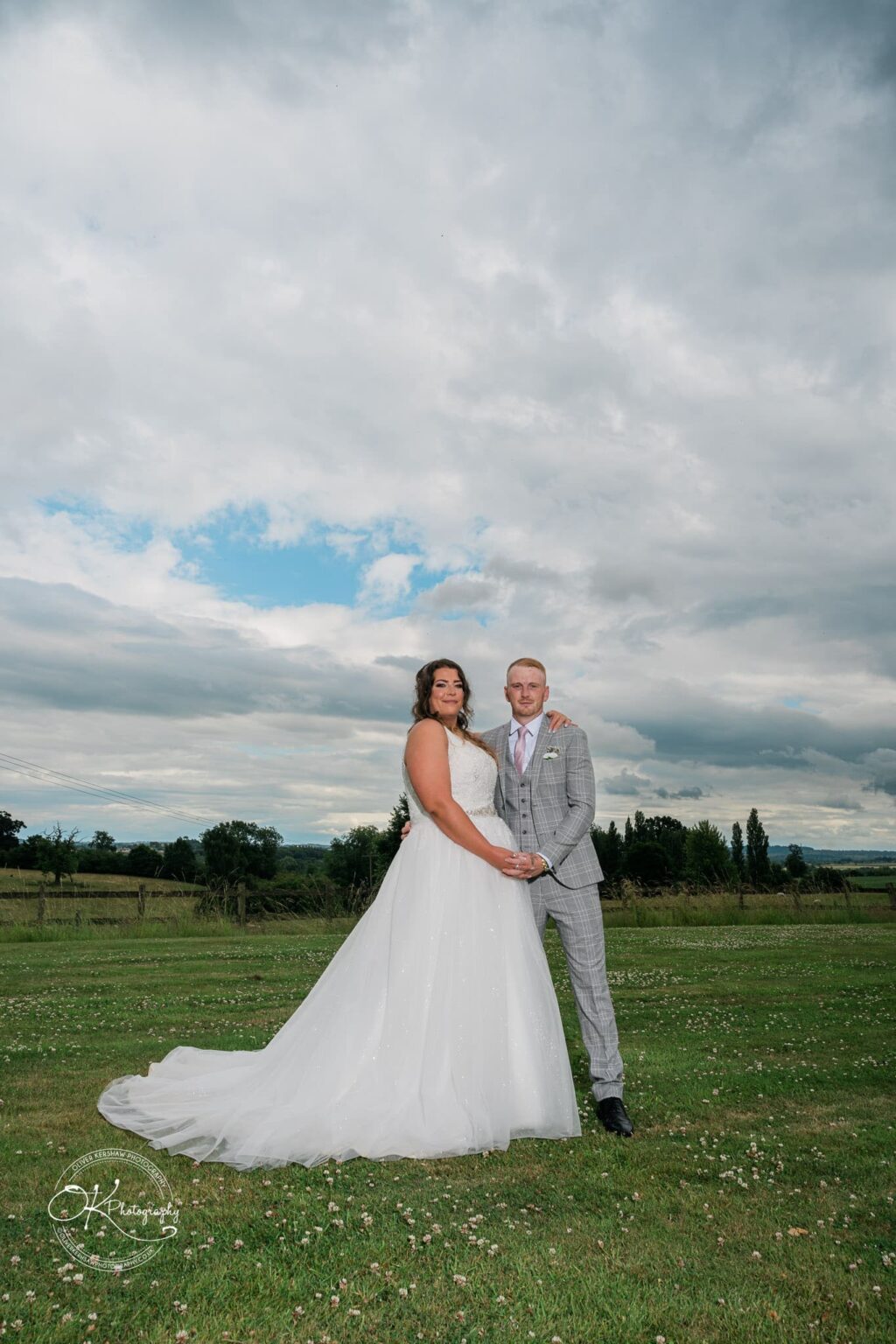 A bride and groom holding hands and posing outdoors on a grassy field with a scenic landscape and cloudy sky in the background.