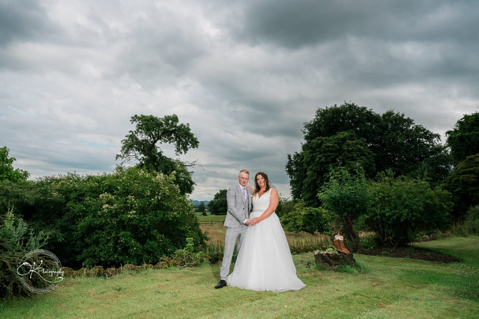 A bride and groom stand together on a grassy area with trees and bushes in the background, under a cloudy sky.