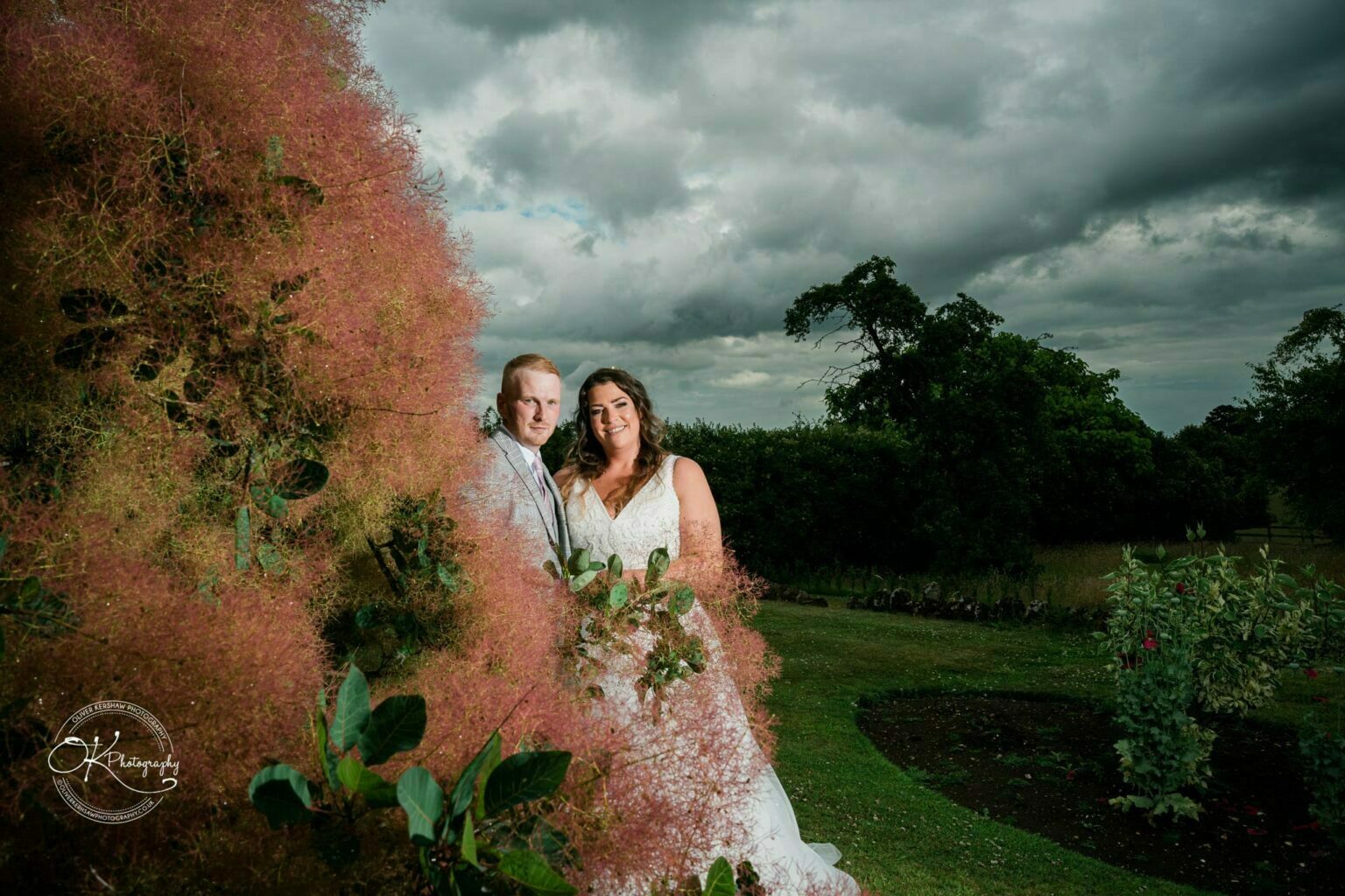 Bride and groom standing beside a tree with pink foliage at Bourton Hall.