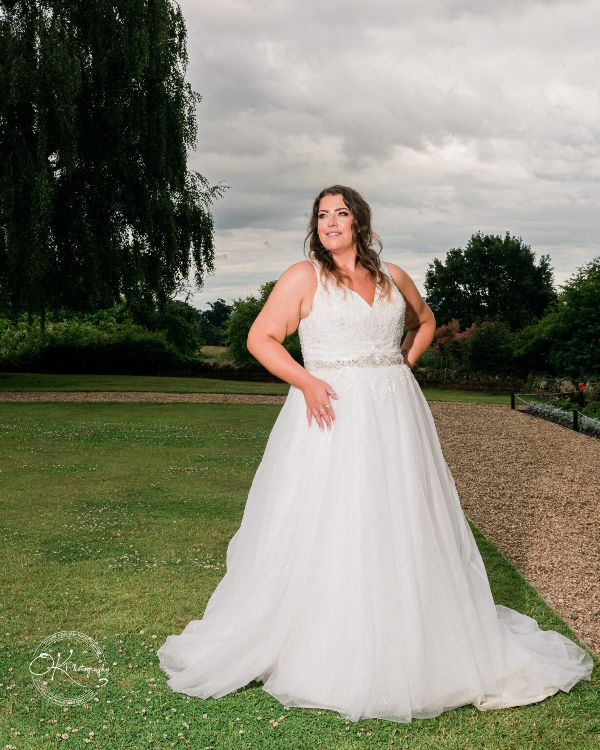 A bride in a white wedding dress posing outdoors on a grassy area with trees in the background.