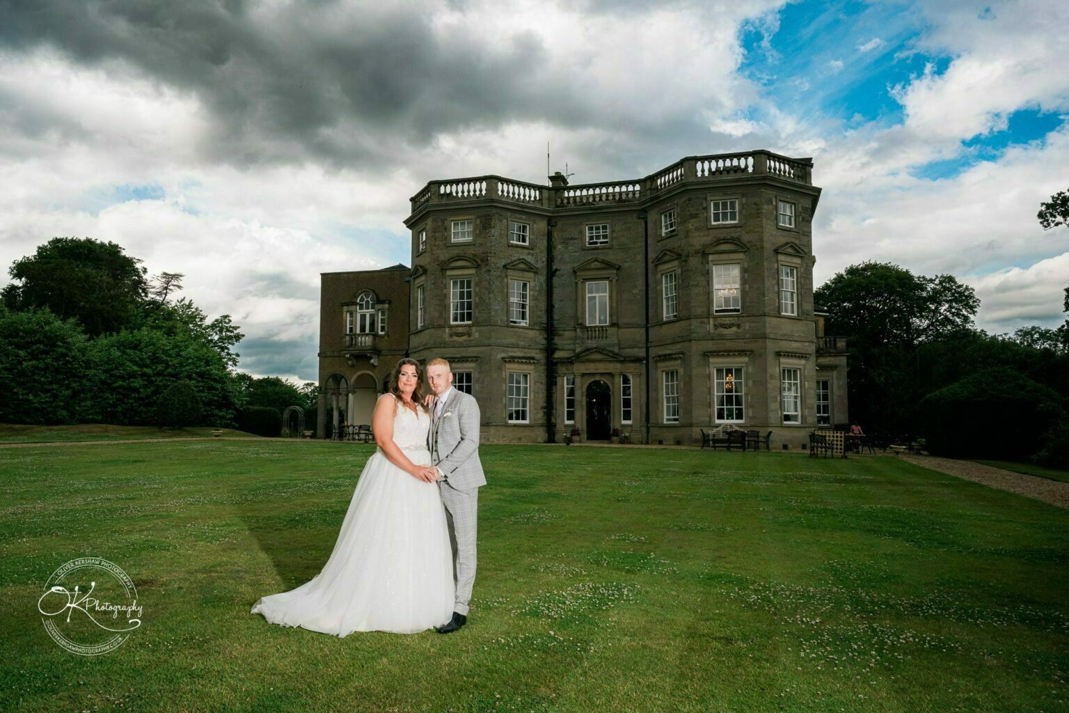 Bride and groom posing on the lawn in front of Bourton Hall under a cloudy sky.