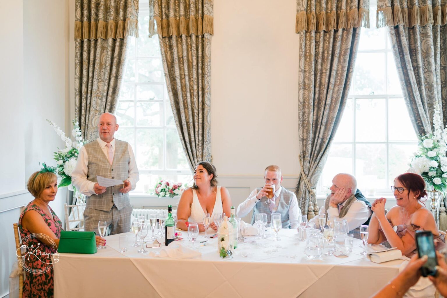 A wedding toast is given by an older man in a grey vest at a formal reception with six people seated at a table in front of large windows with elegant curtains at Bourton Hall.