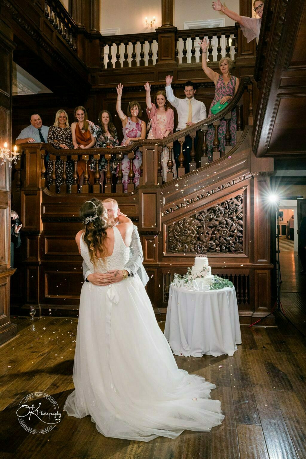 Bride and groom embrace during their first dance, with guests cheering from a wooden staircase and wedding cake on a nearby table, in an elegant venue with intricate woodwork.