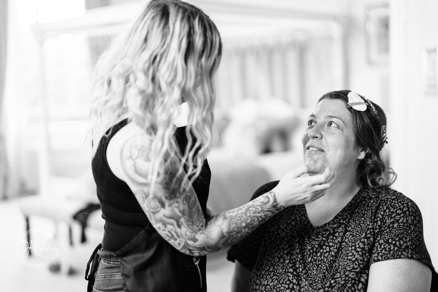 A makeup artist with tattooed arms touching the chin of a smiling woman with a hair clip, preparing for a wedding at Bourton Hall.