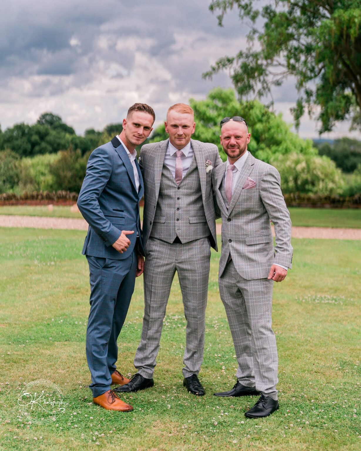 Three men in suits posing on a lawn under a cloudy sky at Bourton Hall.