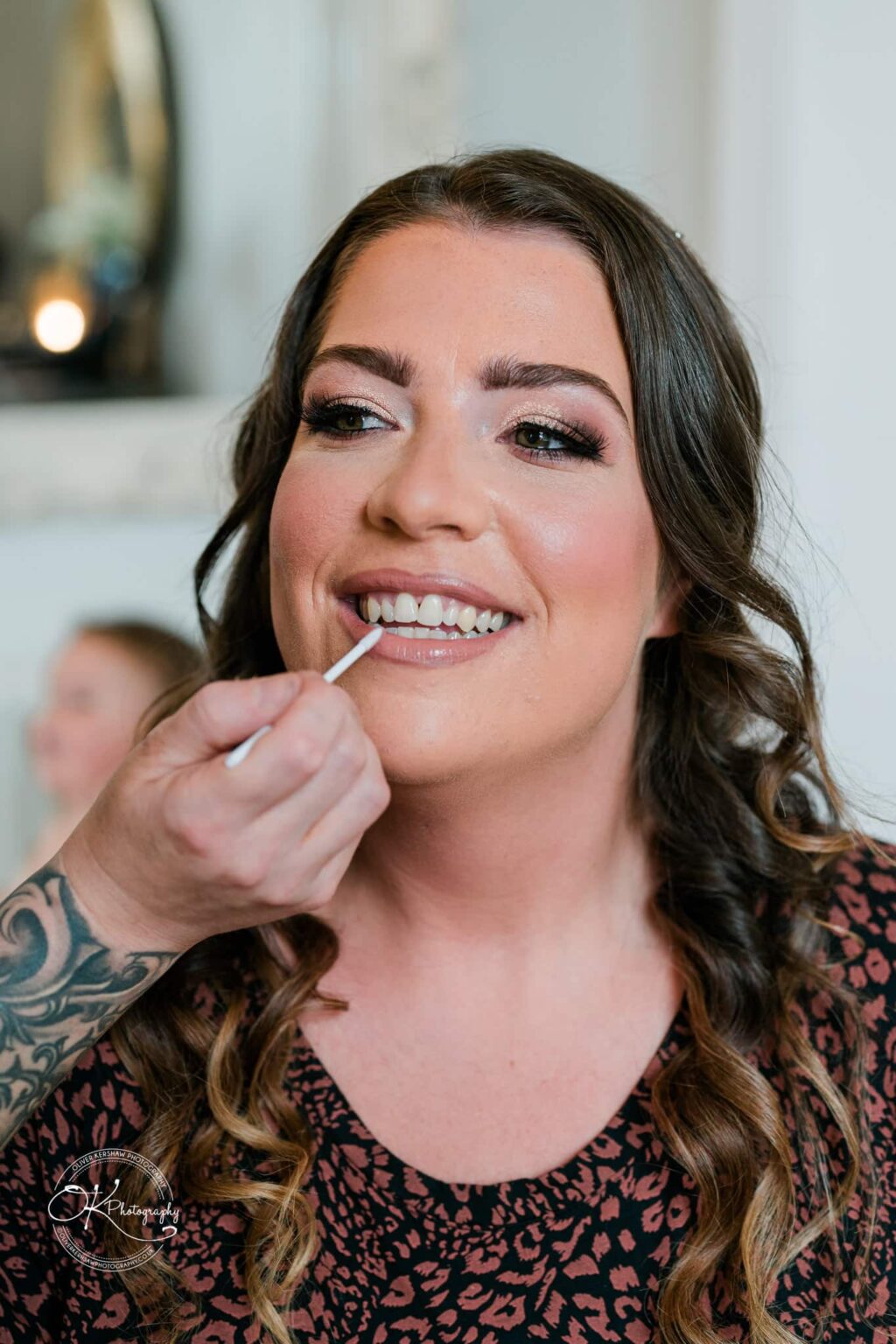 A woman having makeup applied, smiling, with focus on her face and hand applying lip gloss.
