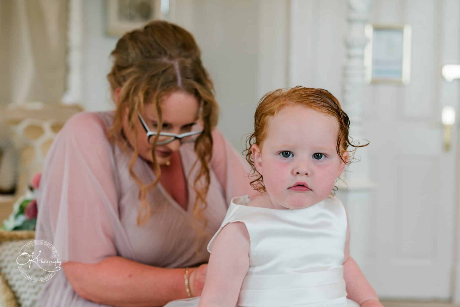 A young child in a white dress with a woman in a soft pink dress and glasses helping her in the background.