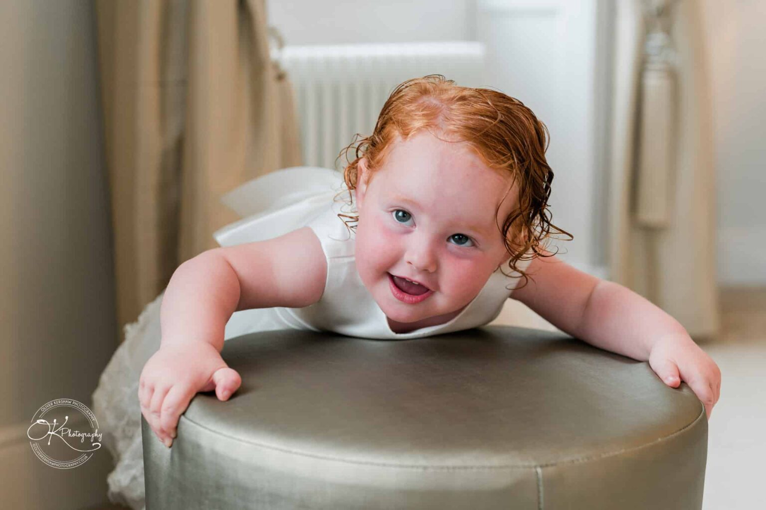 A young child in a white dress leans on a round stool, smiling at the camera.