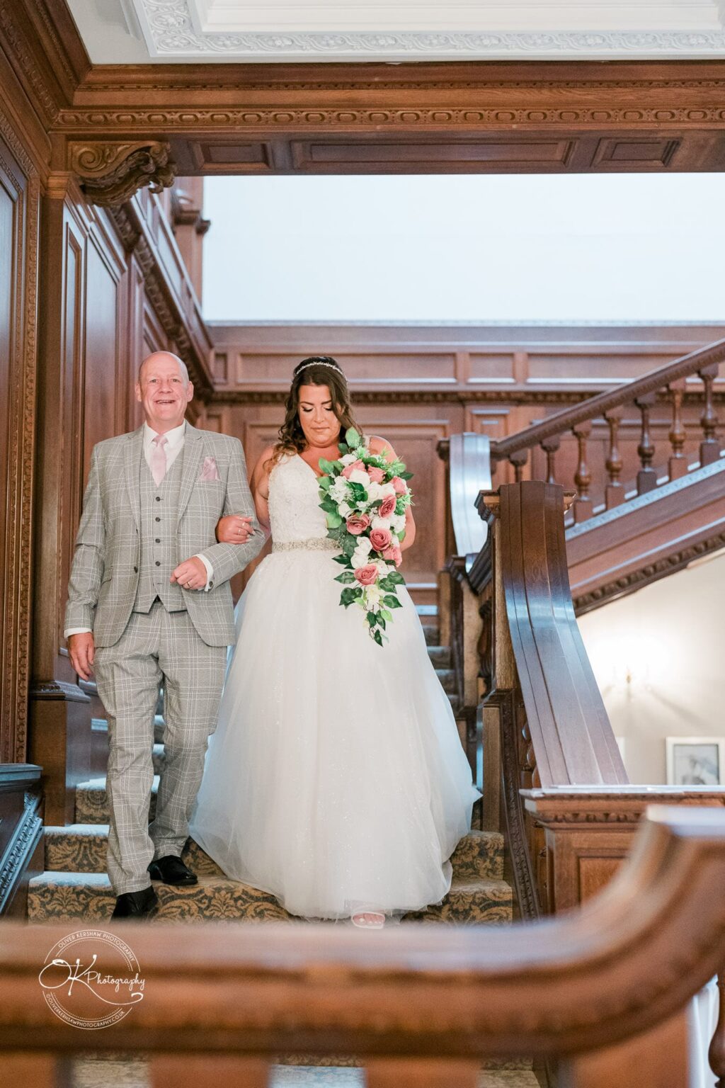 A bride holding a pink and white floral bouquet walks down a wooden staircase arm-in-arm with a man in a light-coloured checked suit.