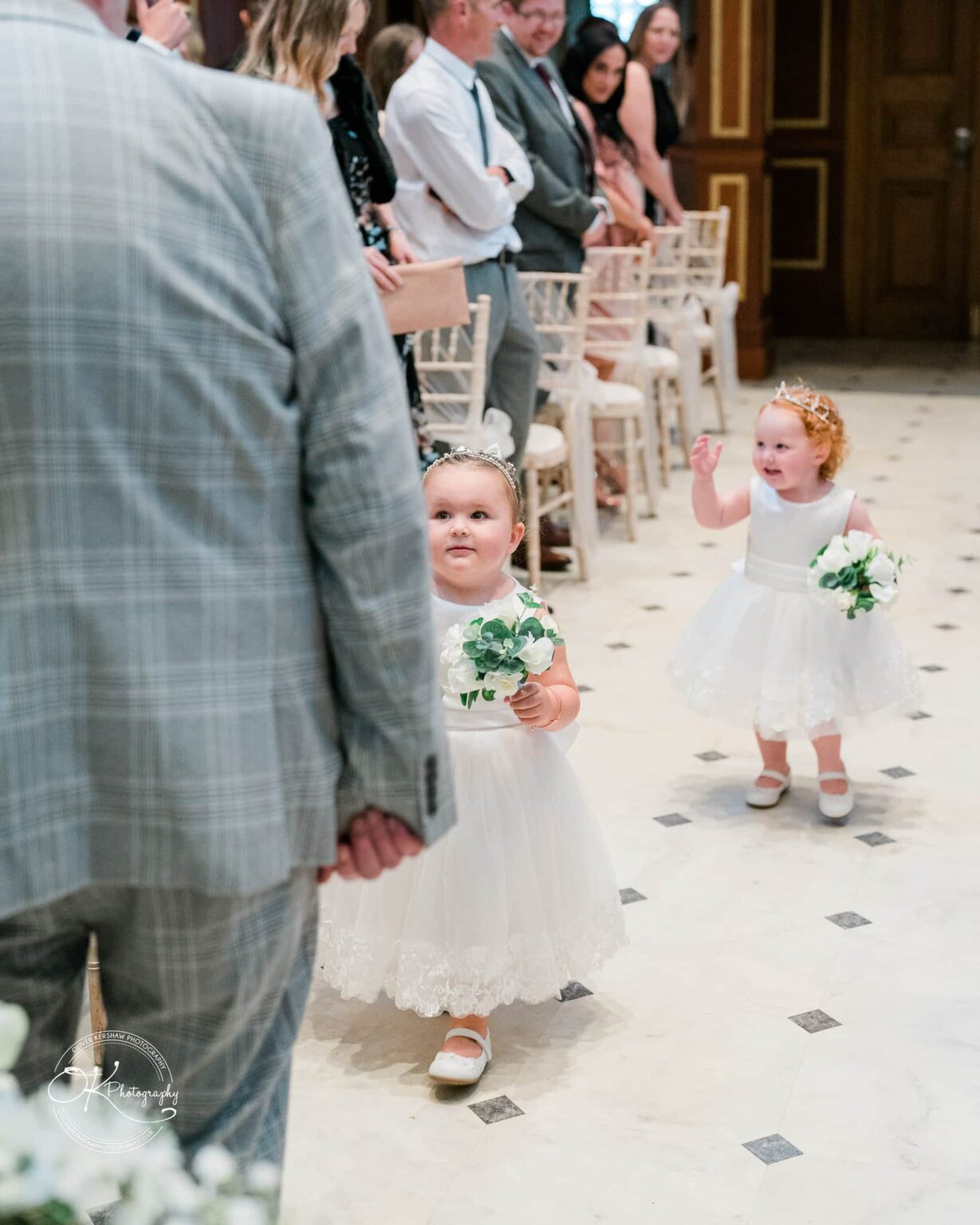 Two young flower girls in white dresses and floral crowns walk down the aisle, carrying small bouquets, surrounded by seated wedding guests.