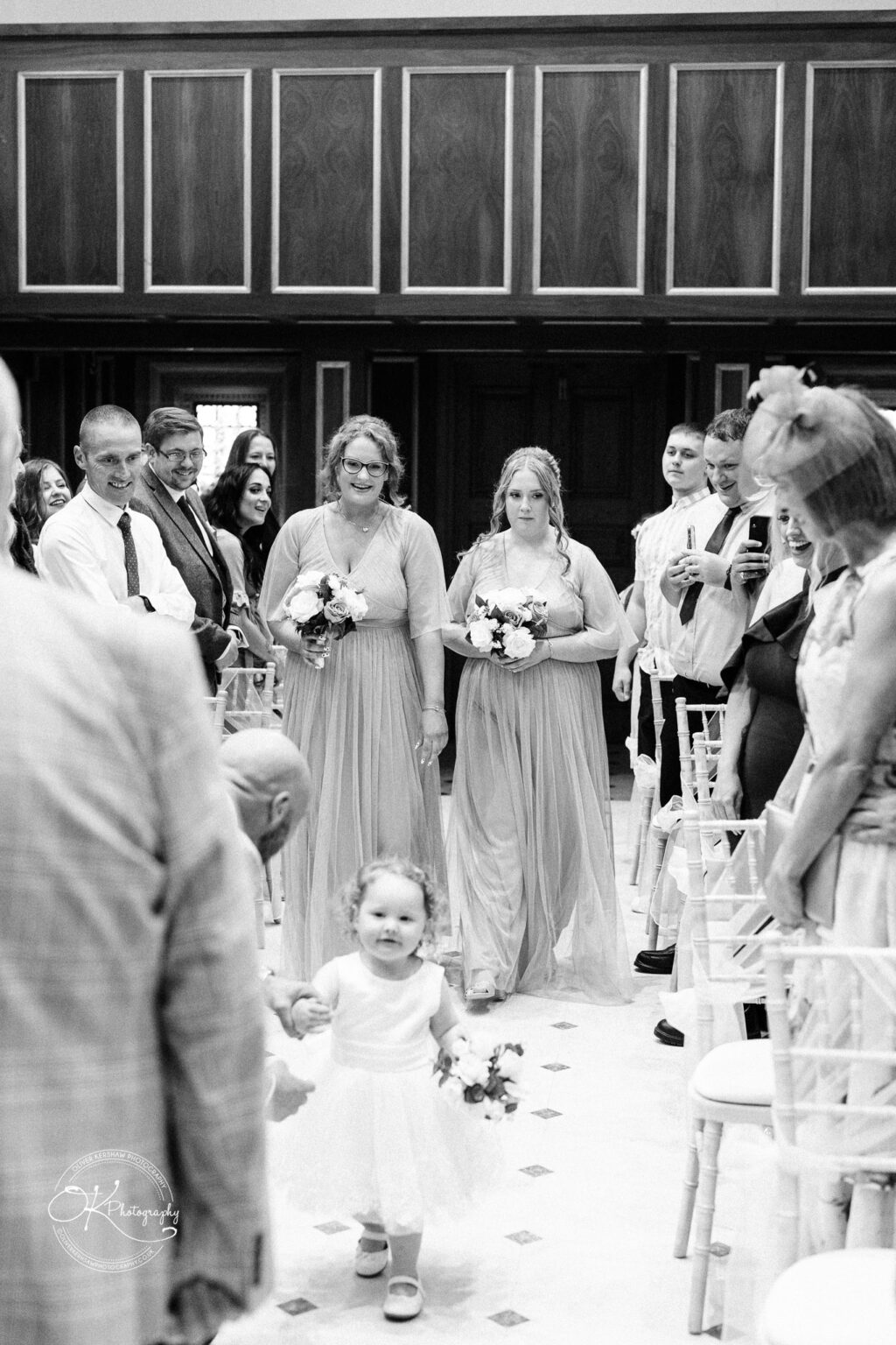 A black and white photo of a wedding procession inside Bourton Hall, featuring a toddler walking with a small bouquet and two bridesmaids in dresses holding bouquets, surrounded by smiling guests.