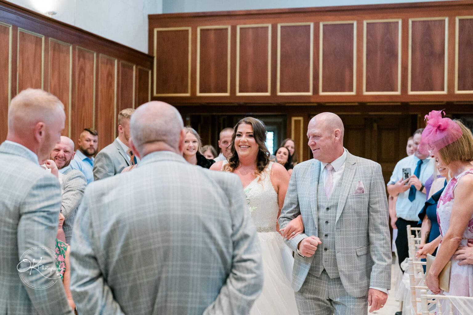 Bride walking down the aisle at Bourton Hall, accompanied by an older man and surrounded by guests.
