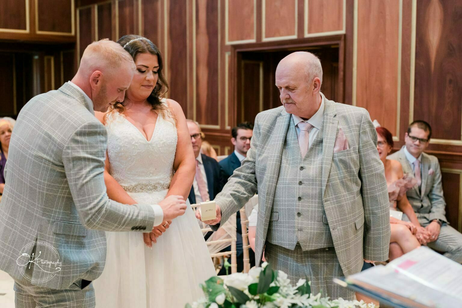 Groom and bride in wedding ceremony with an older man in a plaid suit presenting a small box, surrounded by seated guests in a wood-paneled room.