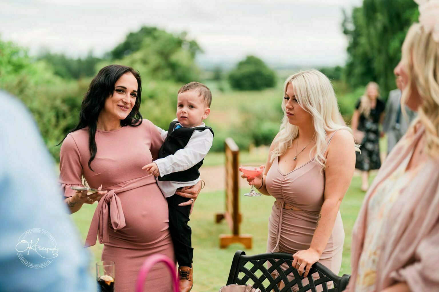 A woman holding a young boy, standing next to another woman holding a drink, with a blurred outdoor background.
