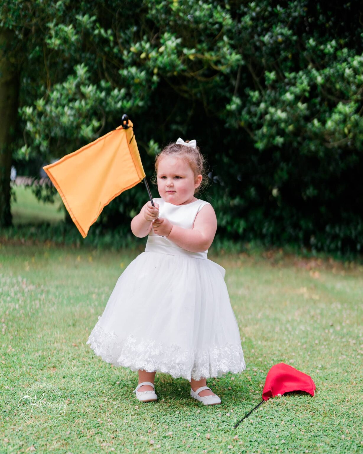 A little girl in a white dress holding an orange flag stands on grass with a red hat on the ground nearby, surrounded by lush greenery.