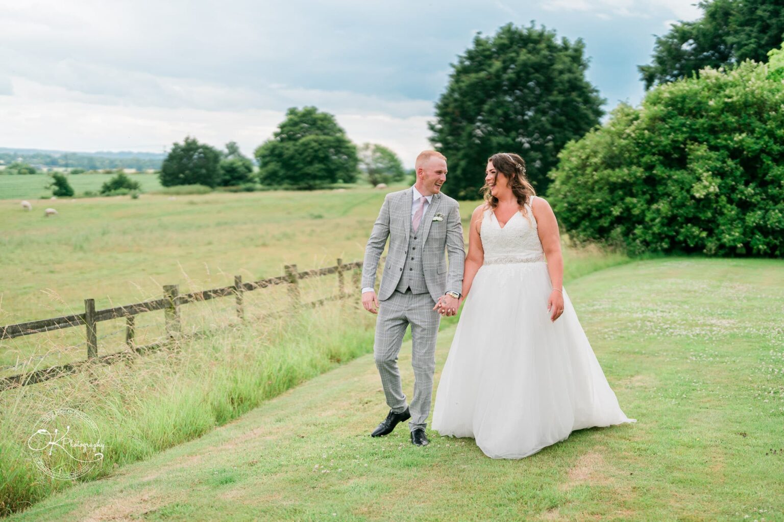 A bride and groom holding hands and smiling while walking on a grassy field with trees and a wooden fence in the background at Bourton Hall.