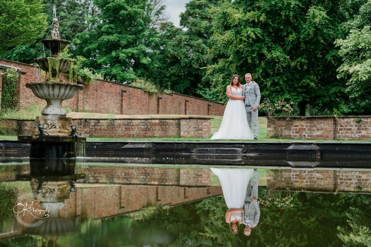 A bride and groom stand by a fountain with their reflections visible in the water at Bourton Hall.