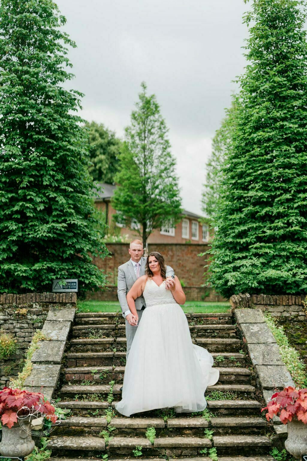 A couple standing on stone steps surrounded by greenery, with the bride in a white dress and the groom in a grey suit, at Bourton Hall.