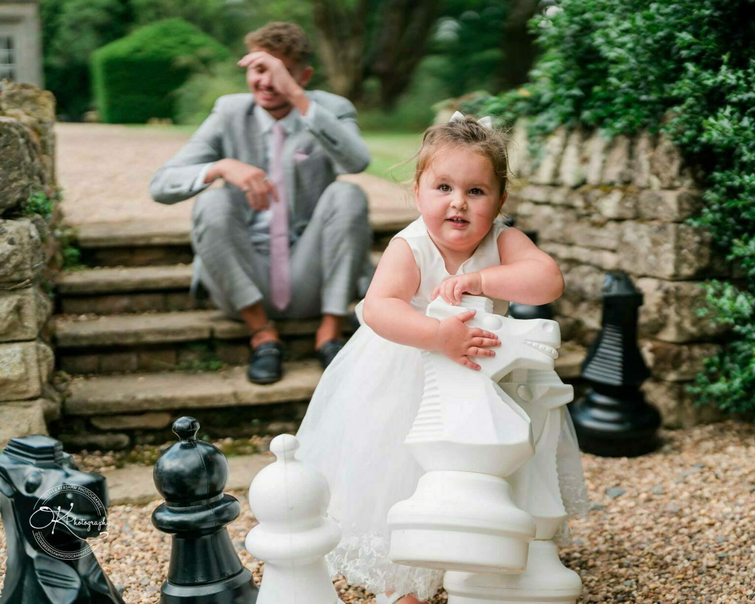 A toddler in a white dress holds a large white chess piece, with a man in a grey suit sitting in the background.