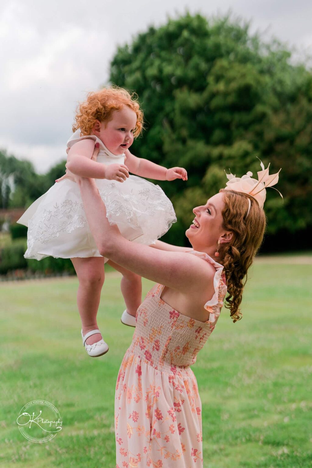 A woman in a floral dress lifts a baby in a white dress and shoes, both smiling and outdoors with greenery in the background.