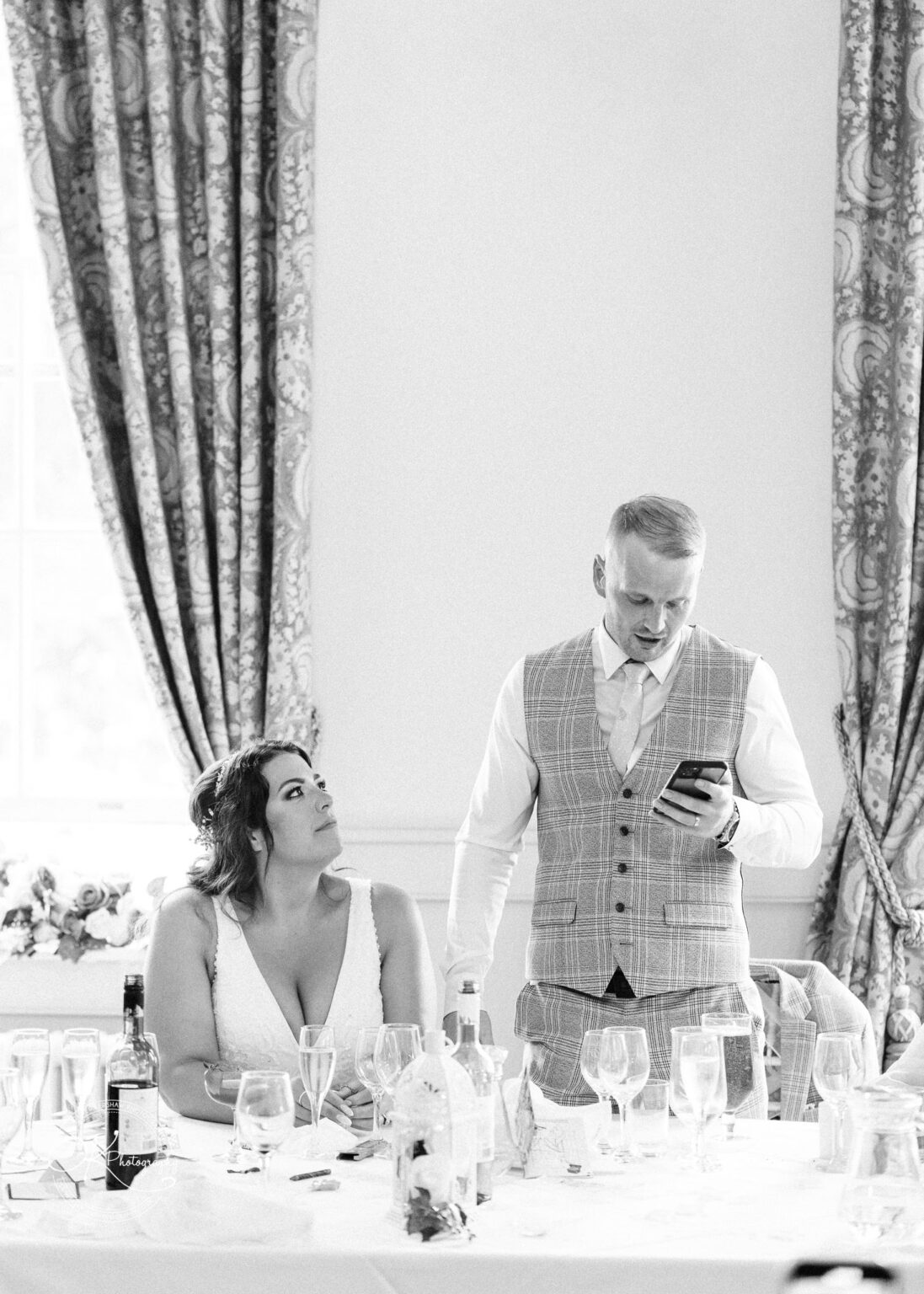 A bride sitting and looking up at the groom who is standing and reading from his phone during a wedding reception speech.