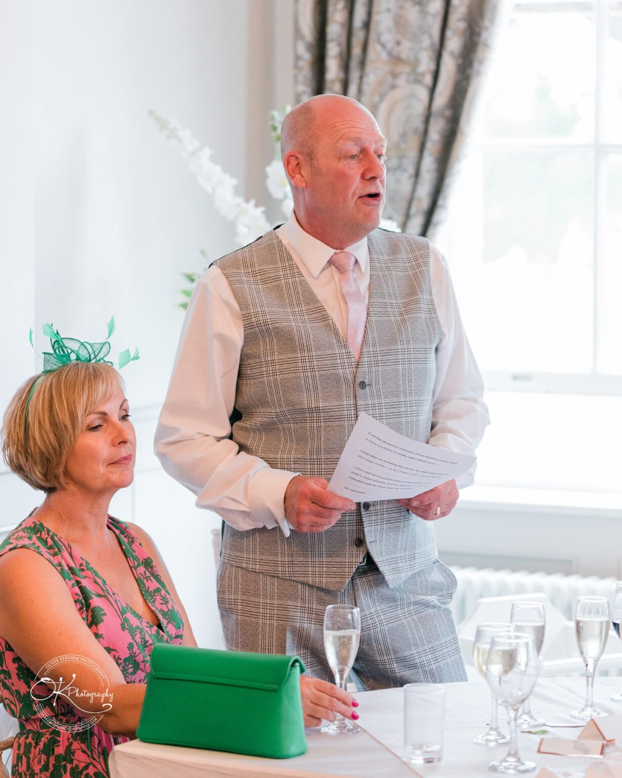 A man in a grey plaid waistcoat and pink tie giving a speech at a wedding reception, standing next to a seated woman in a floral dress with a green fascinator.