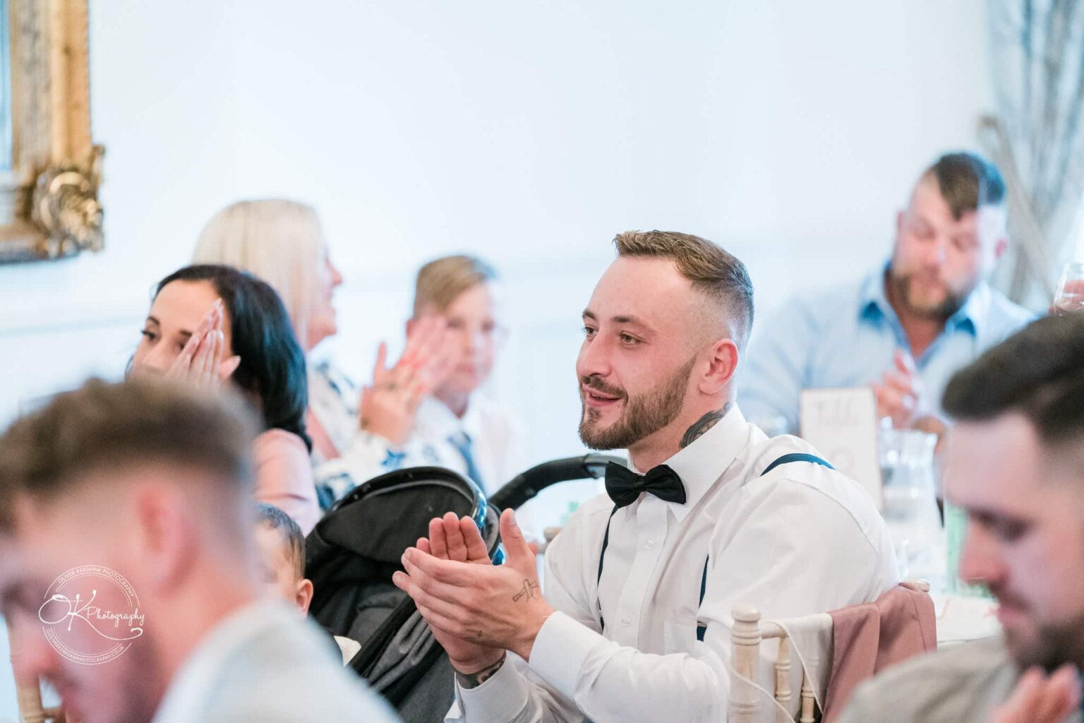 A man dressed in formal attire clapping at a wedding reception, with other guests in the background.
