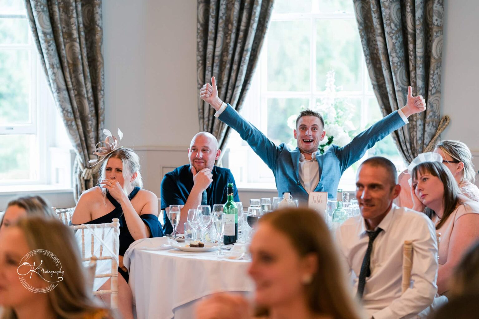 Guests sitting at a table at a wedding reception, with one man in a blue suit excitedly giving two thumbs up.