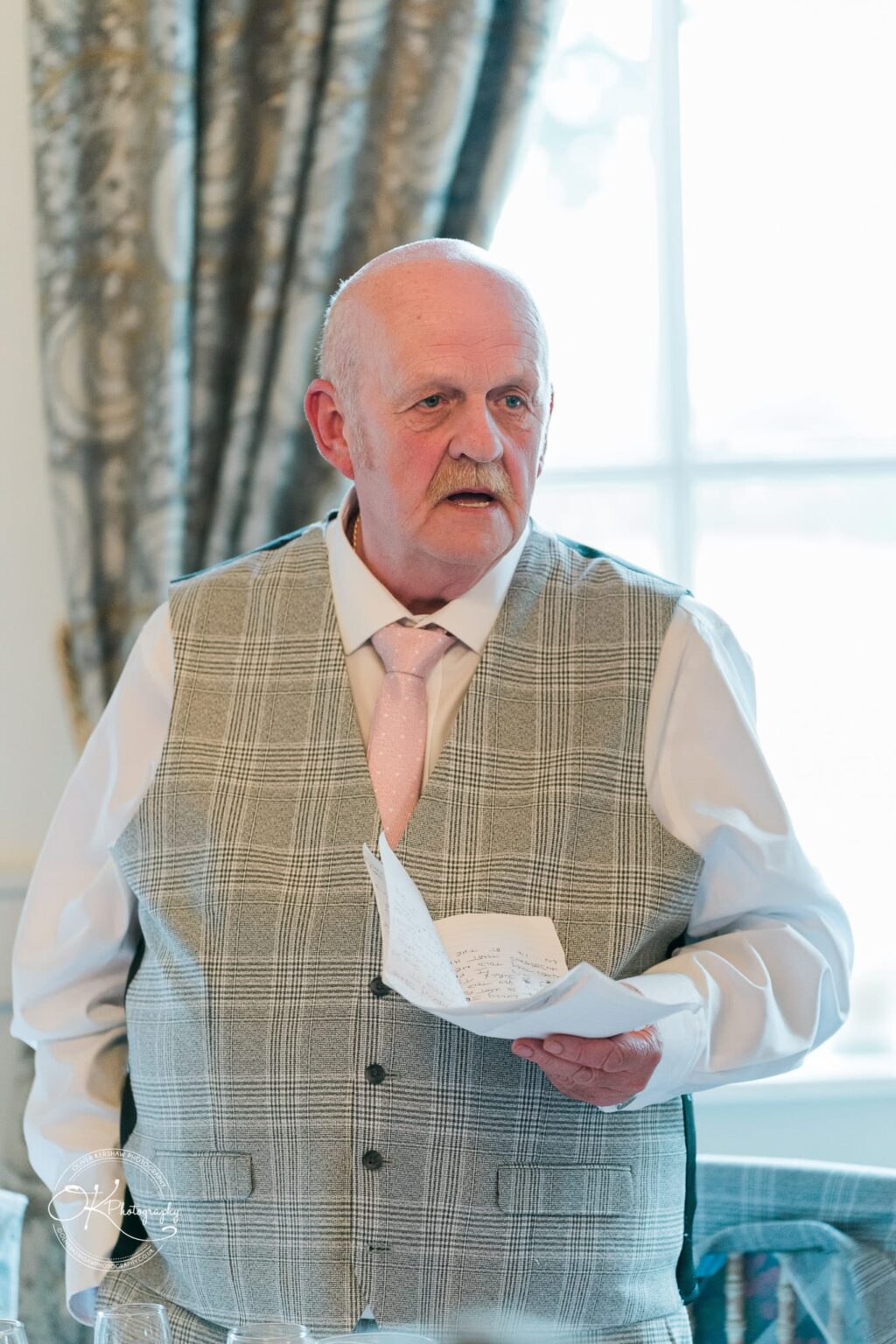 An older man in a grey checked waistcoat and pink tie giving a speech at a wedding.