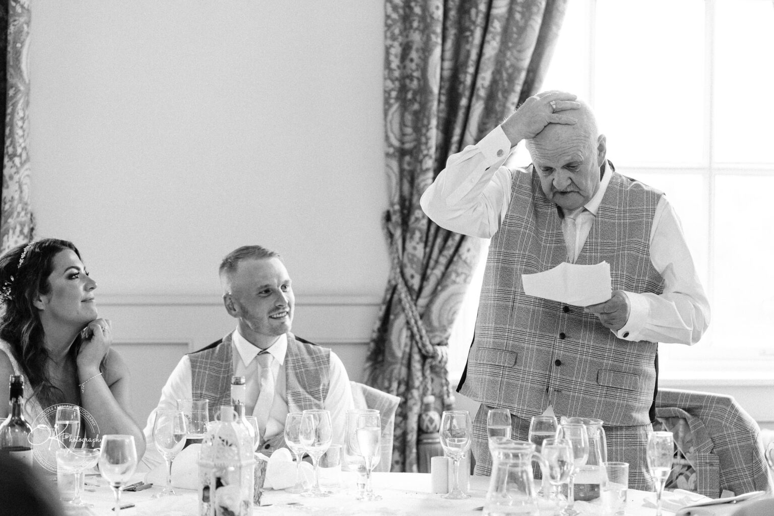An elderly man giving a speech at a wedding reception while a couple looks on, with an elegantly decorated window in the background.
