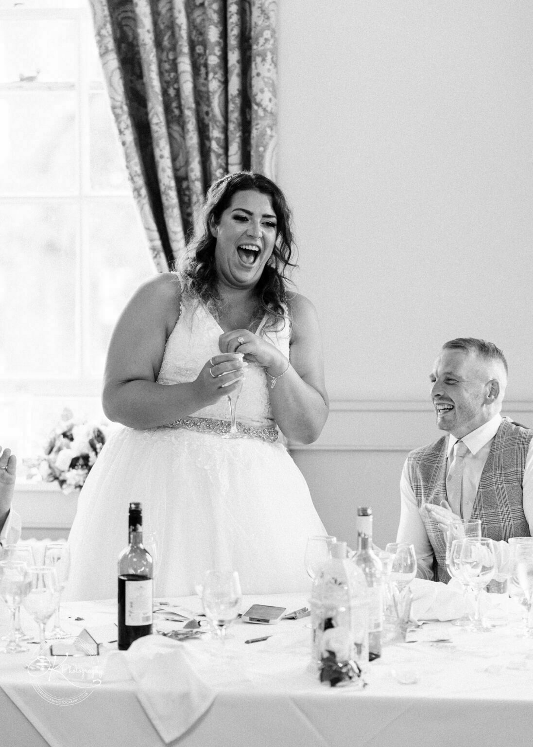 Bride laughing and holding a champagne flute while standing beside a seated man during a wedding reception.
