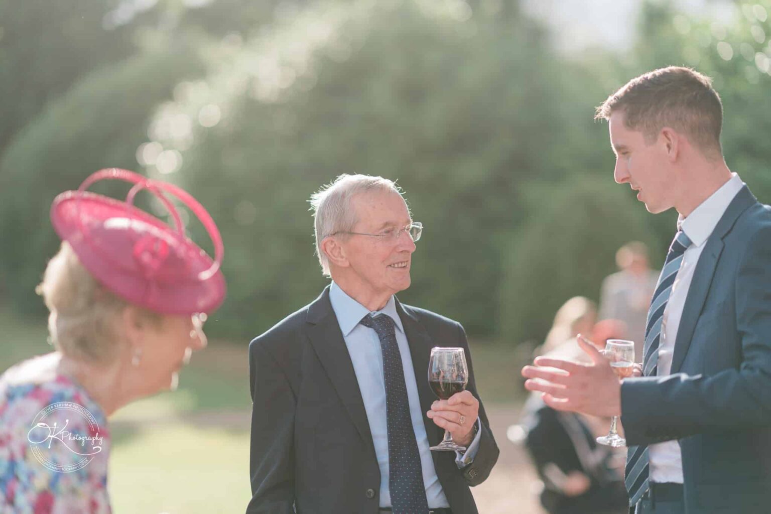 Three people in formal attire conversing outside, one woman wearing a pink fascinator, one older man holding a glass of wine, and one younger man gesturing.