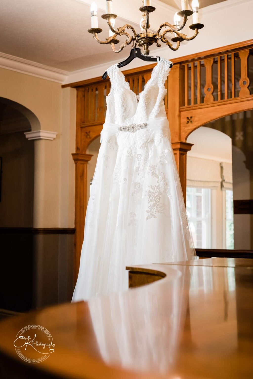 Brooksby Hall Wedding Photography White lace wedding dress hanging from a chandelier in a room with wooden decor and arched doorways.