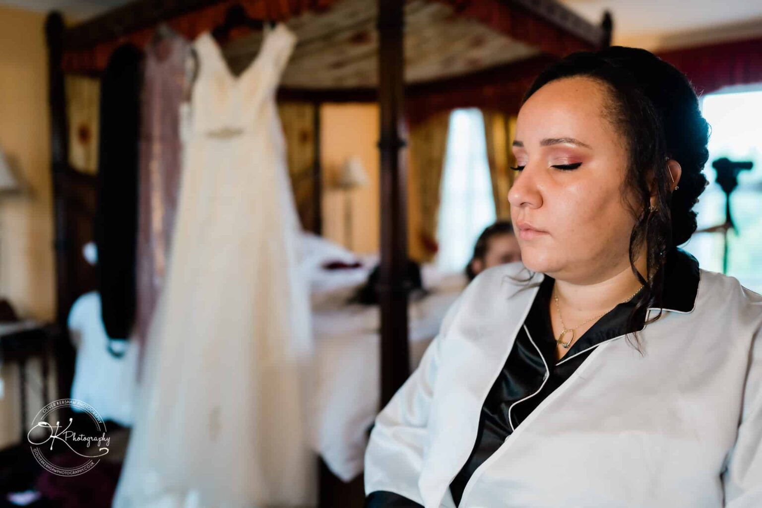 Brooksby Hall Wedding Photography A woman in a white robe with her eyes closed in a room with a wedding dress hanging in the background.