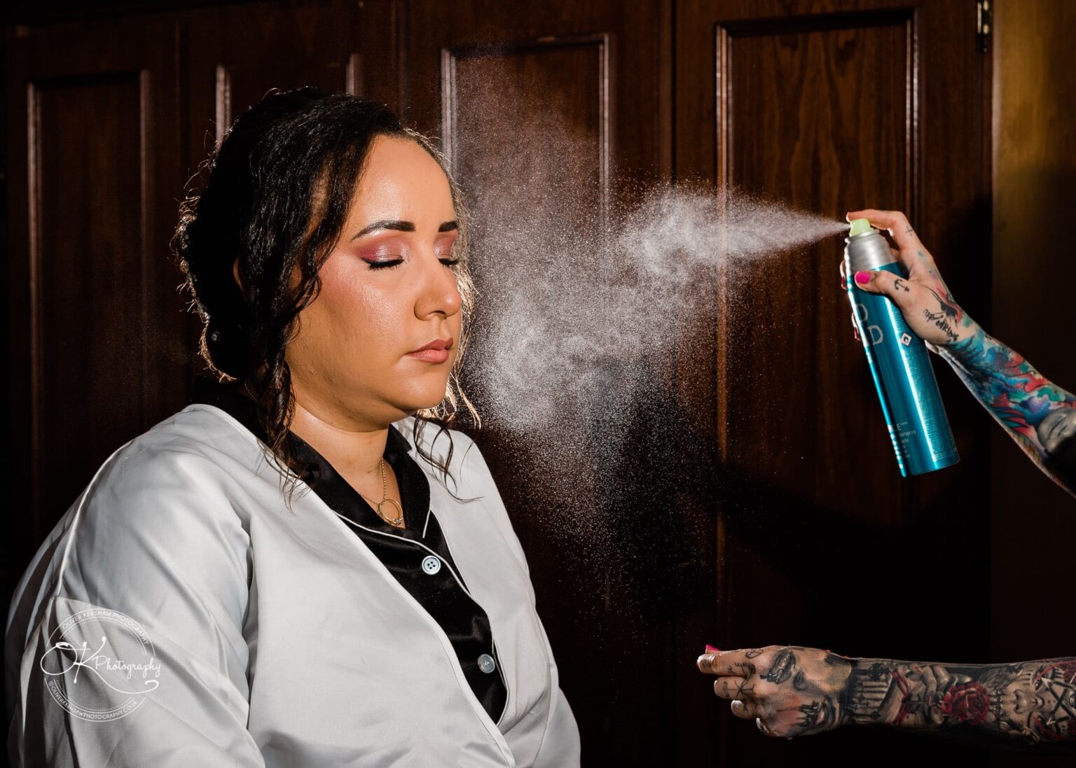 Brooksby Hall Wedding Photography A woman with closed eyes being sprayed with hairspray by a tattooed individual.