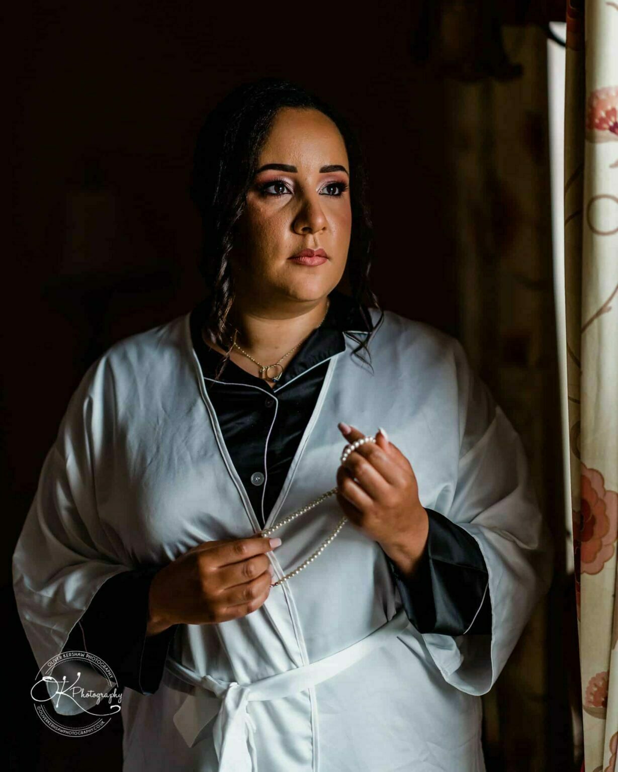 Brooksby Hall Wedding Photography Woman in a robe holding a pearl necklace, gazing out of a window.